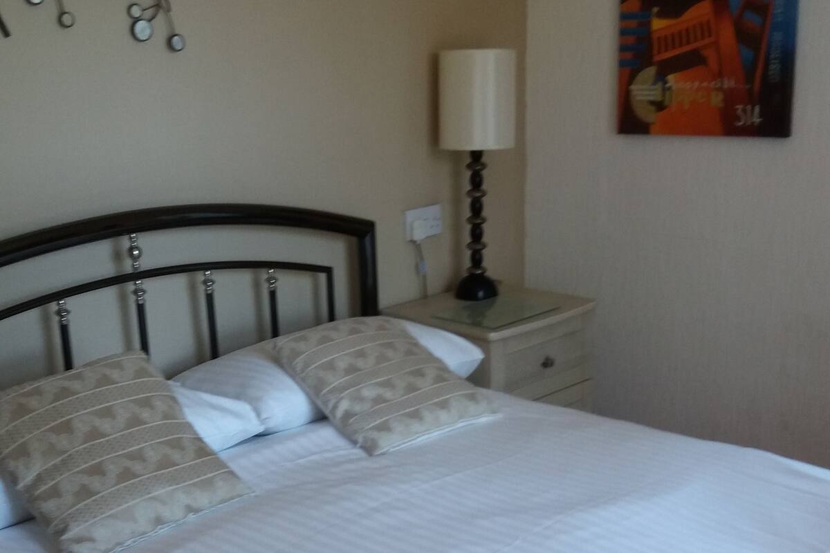 A neatly made bed with white linens and patterned pillows, a bedside lamp, a wall clock, and a framed picture on the wall.