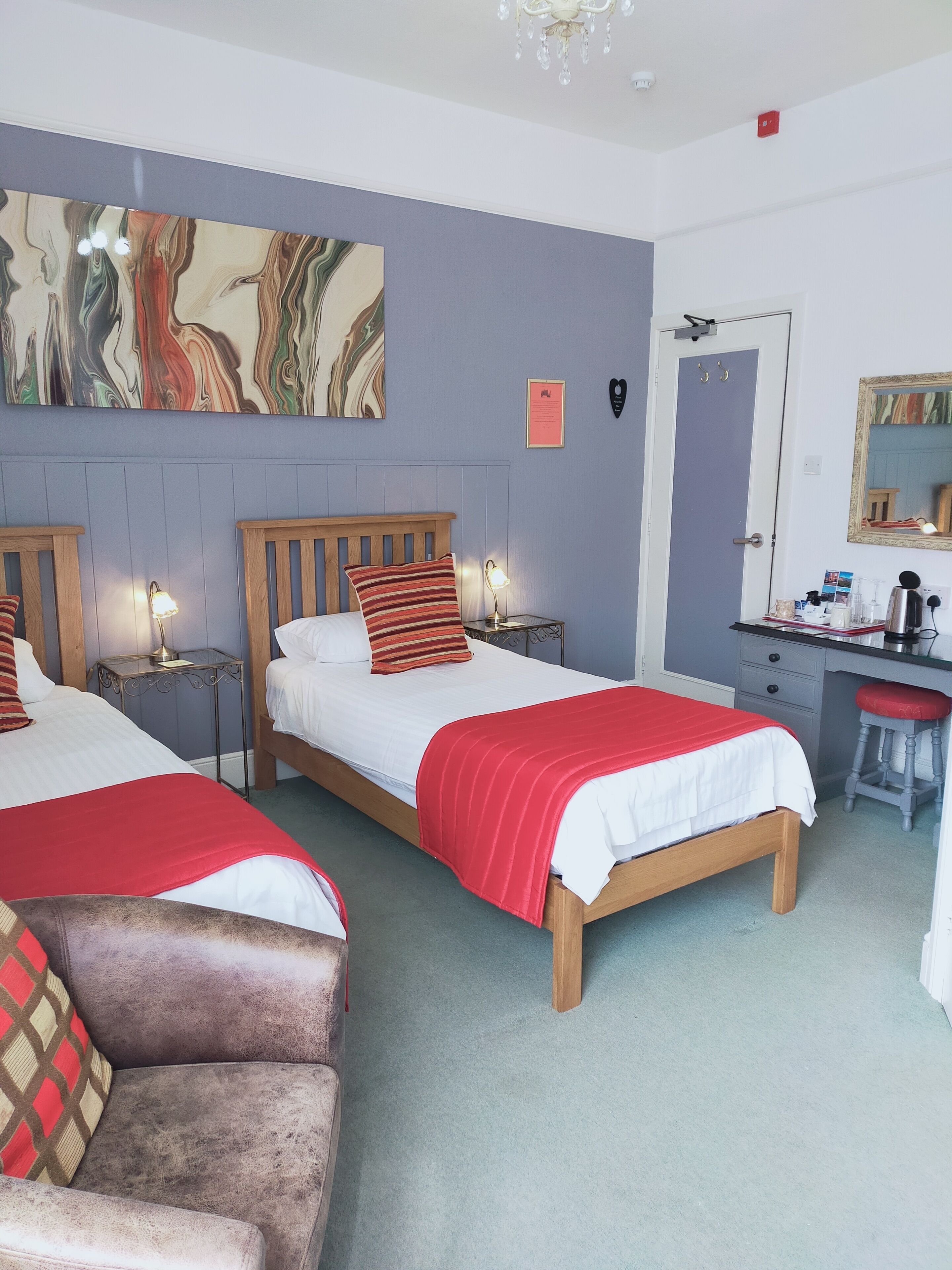 Habitación de hotel con dos camas de madera, cada una con cubrecama rojo y blanco, un cuadro abstracto grande en la pared, un escritorio con una silla y un espejo.