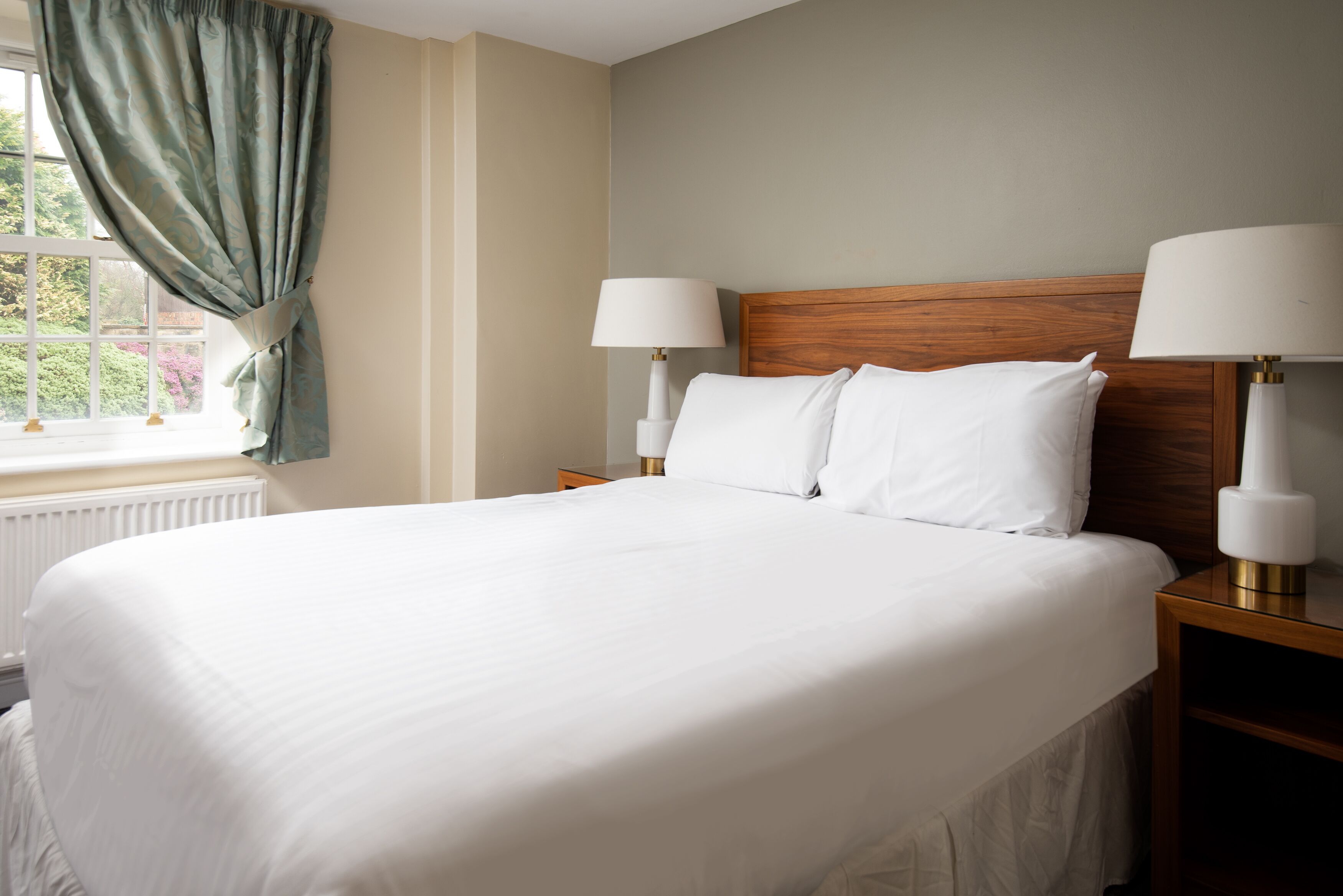 A neatly made bed with white linens, two bedside lamps, and a view of greenery through a window with curtains.