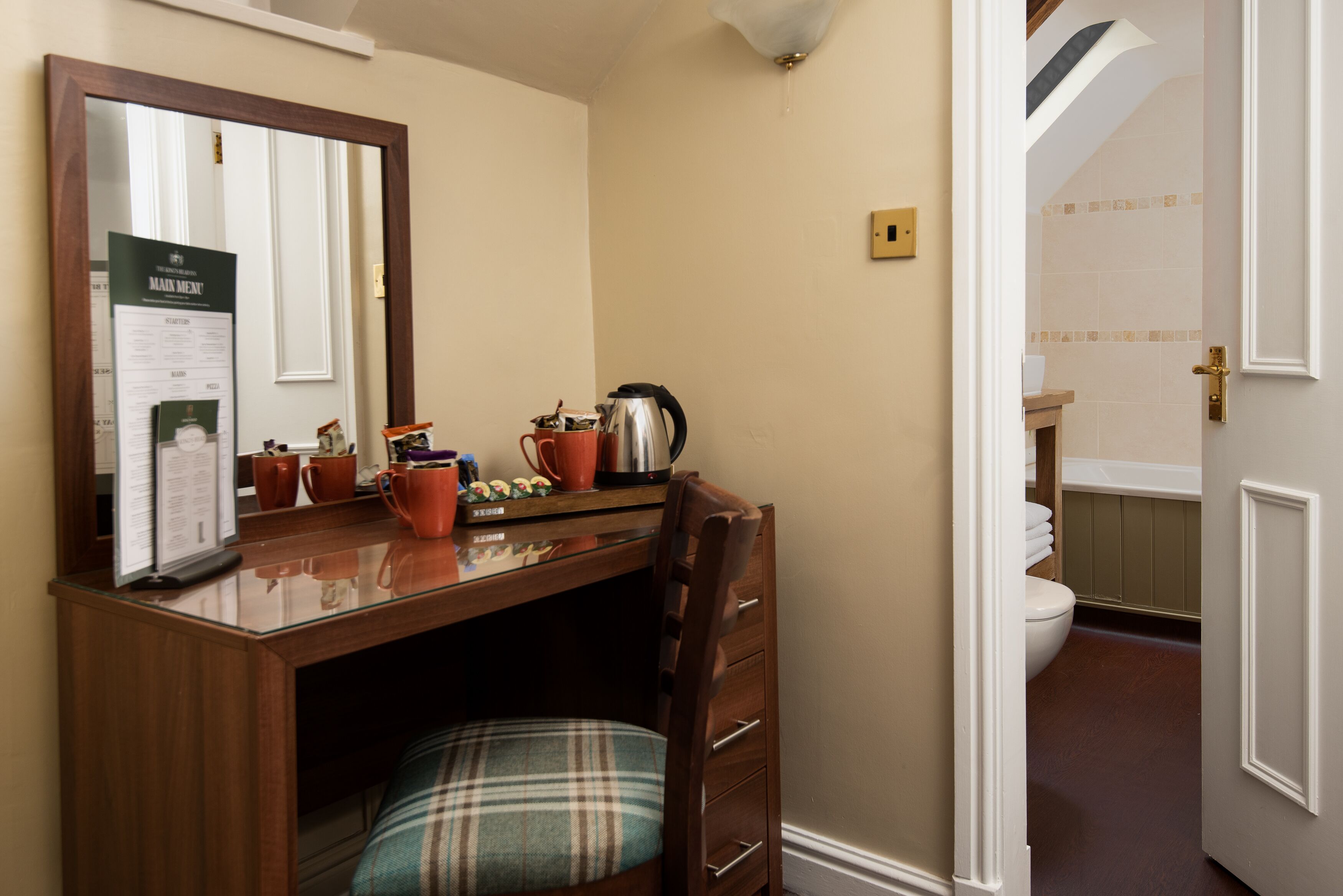 A compact room with a wooden desk, a chair, a mirror, and a small kitchenette area.