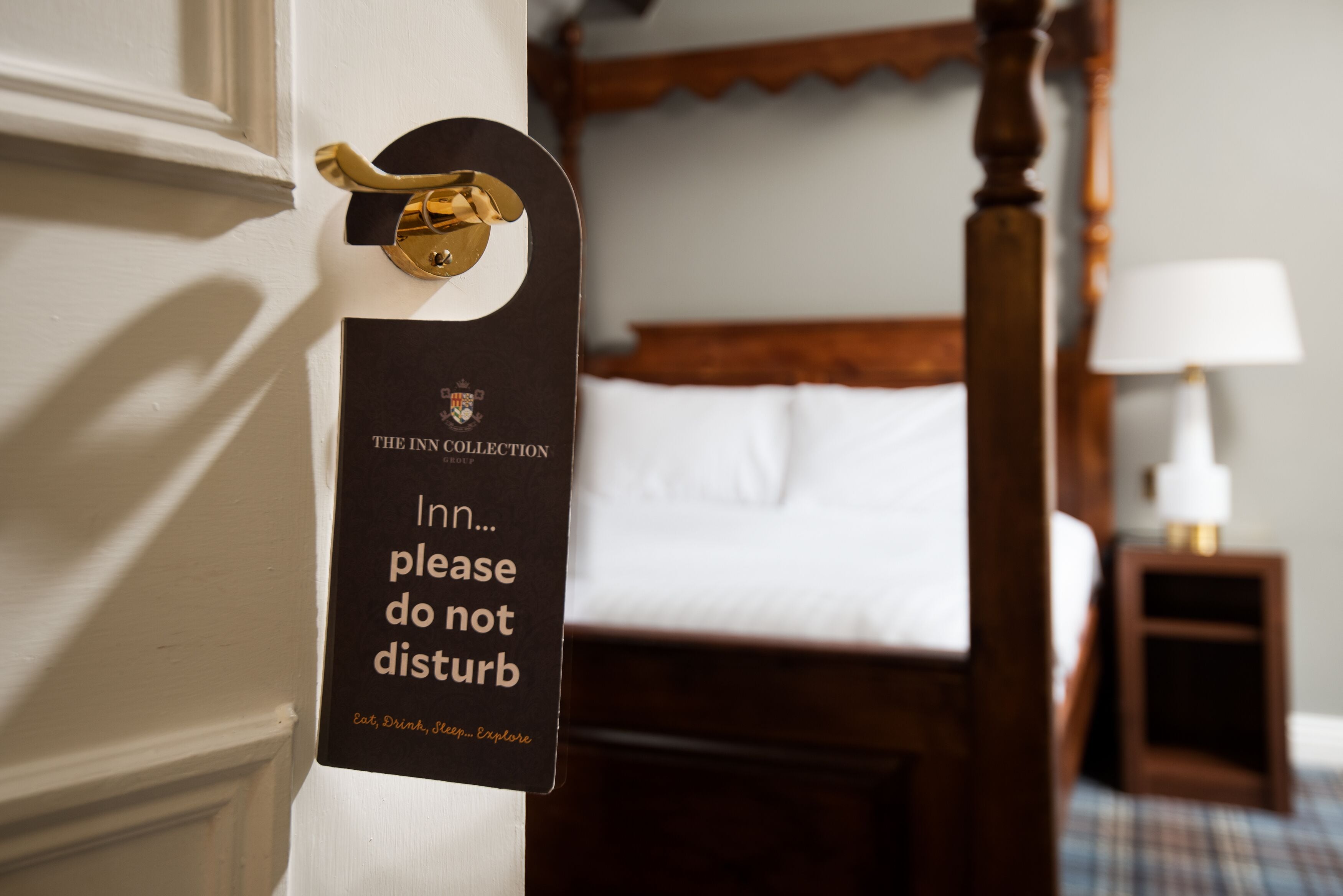 A hotel room with a bed, a bedside table with a lamp, and a sign on the door.