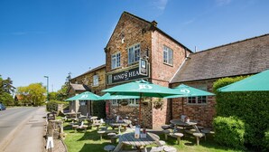 A traditional brick building with a sign reading "King's Head Inn." It features outdoor wooden tables and green umbrellas, with a clear sky above.