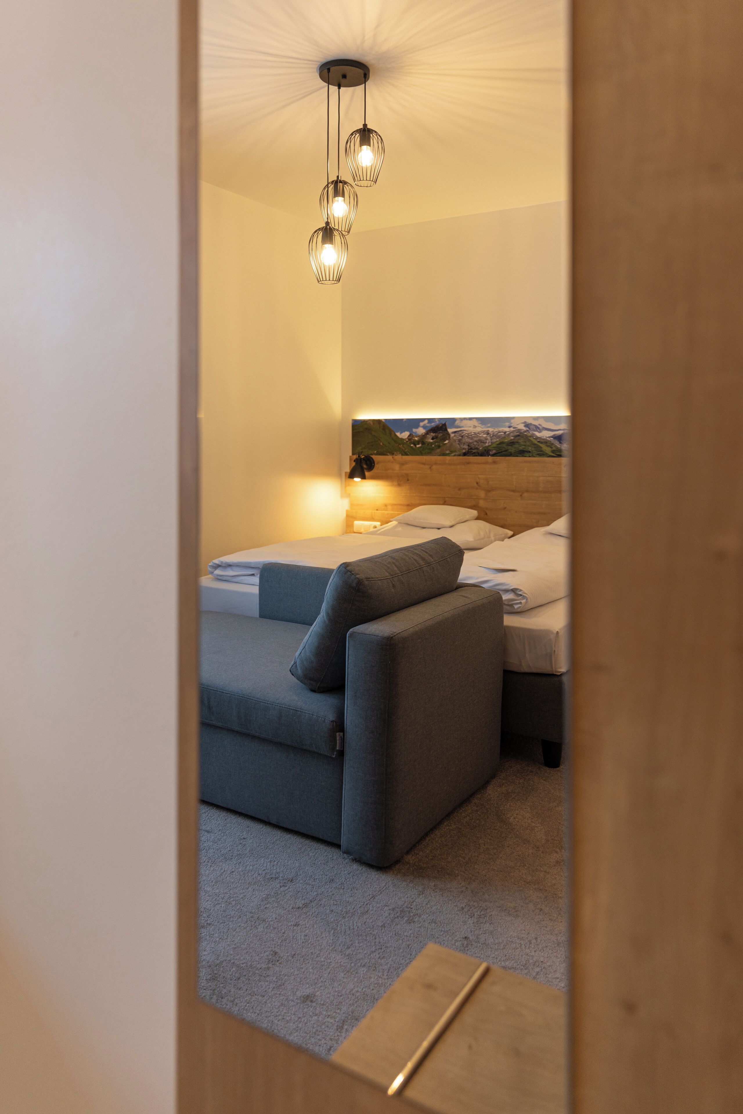 Ein Schlafzimmer mit Bett, grauem Sofa, Holz-Kopfteil und Deckenleuchte.