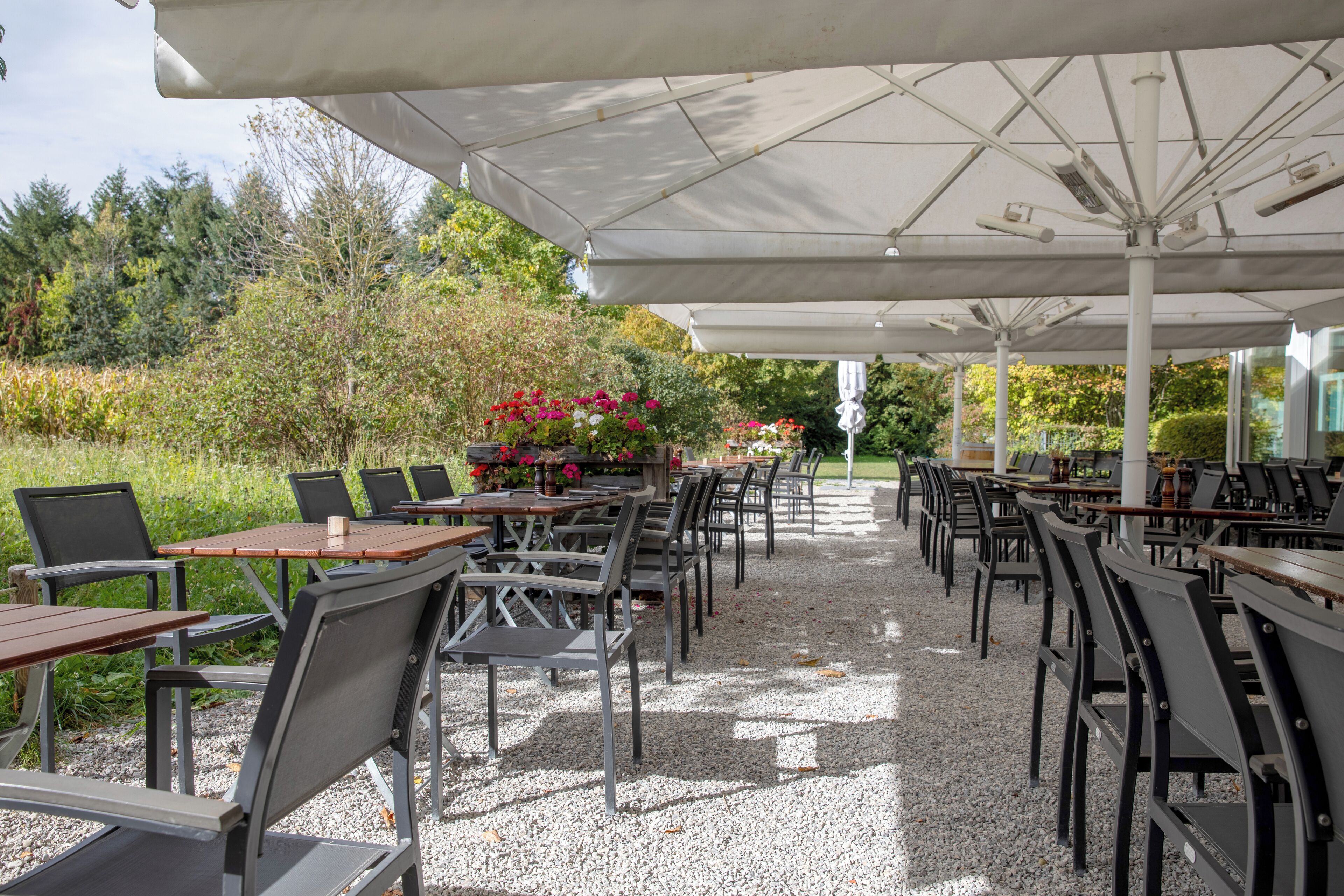 An outdoor patio with tables and chairs under umbrellas, surrounded by greenery and flowers.
