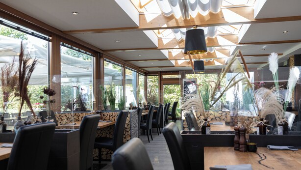 An indoor dining area with wooden tables and black chairs, large windows, and potted plants. - MEAT & STAY HOTEL (Haar)