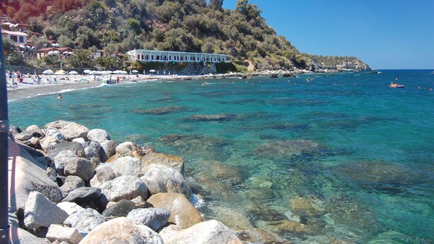 A coastal beach with clear water, rocky shore, and a building with a white facade. - Villaggio Cirucco (Milazzo)