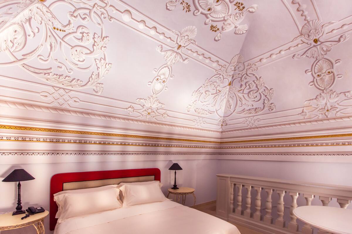 A room with a red headboard, a bed with white linens, a round table, and a small round table with a lamp.