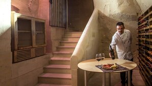 Un chef sirviendo vino en una copa sobre una mesa en una bodega de paredes de piedra, con una estantería llena de vino al fondo.