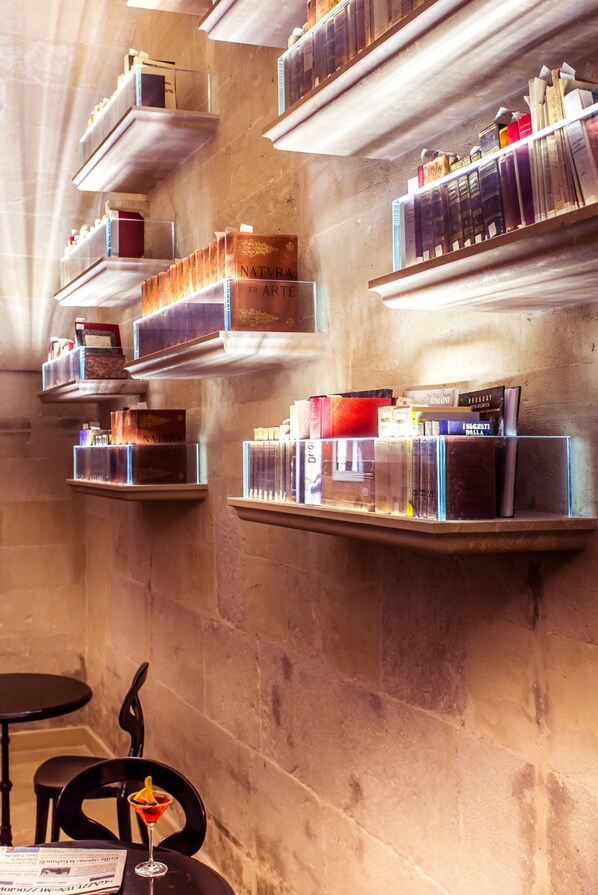 A library with bookshelves against a stone wall, a table with chairs, and a drink on the table. - Palazzo Gattini Luxury Hotel (Matera)