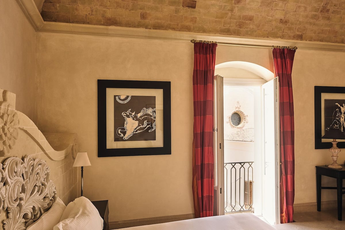A hotel room with a bed, a headboard, a framed picture, a lamp, and a view of the outside.