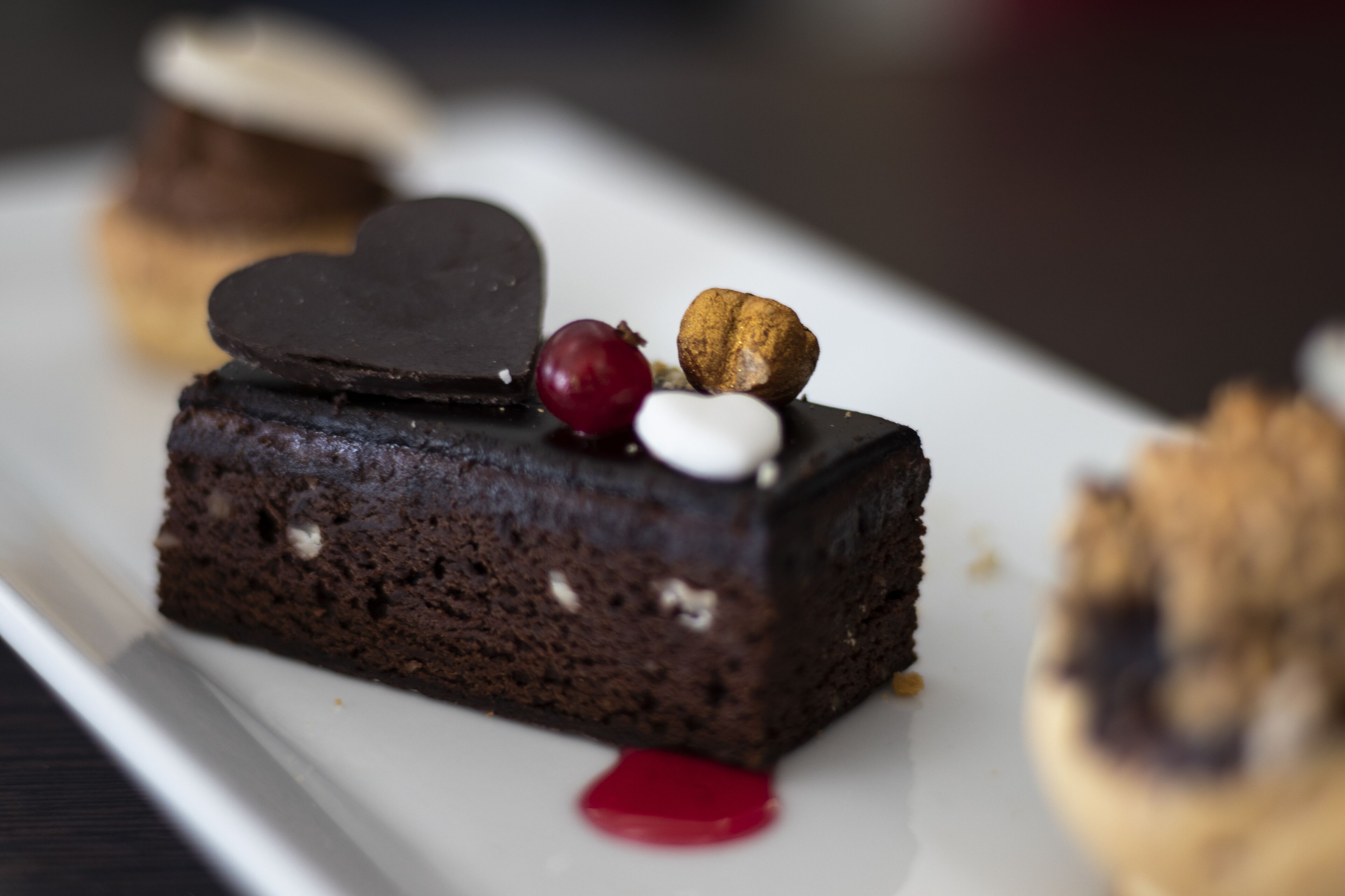 A dessert with a heart-shaped chocolate piece, a red berry, and a nut on a white plate.