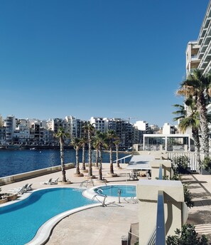 Zona de piscina en un resort, con vista al mar y edificios al fondo.