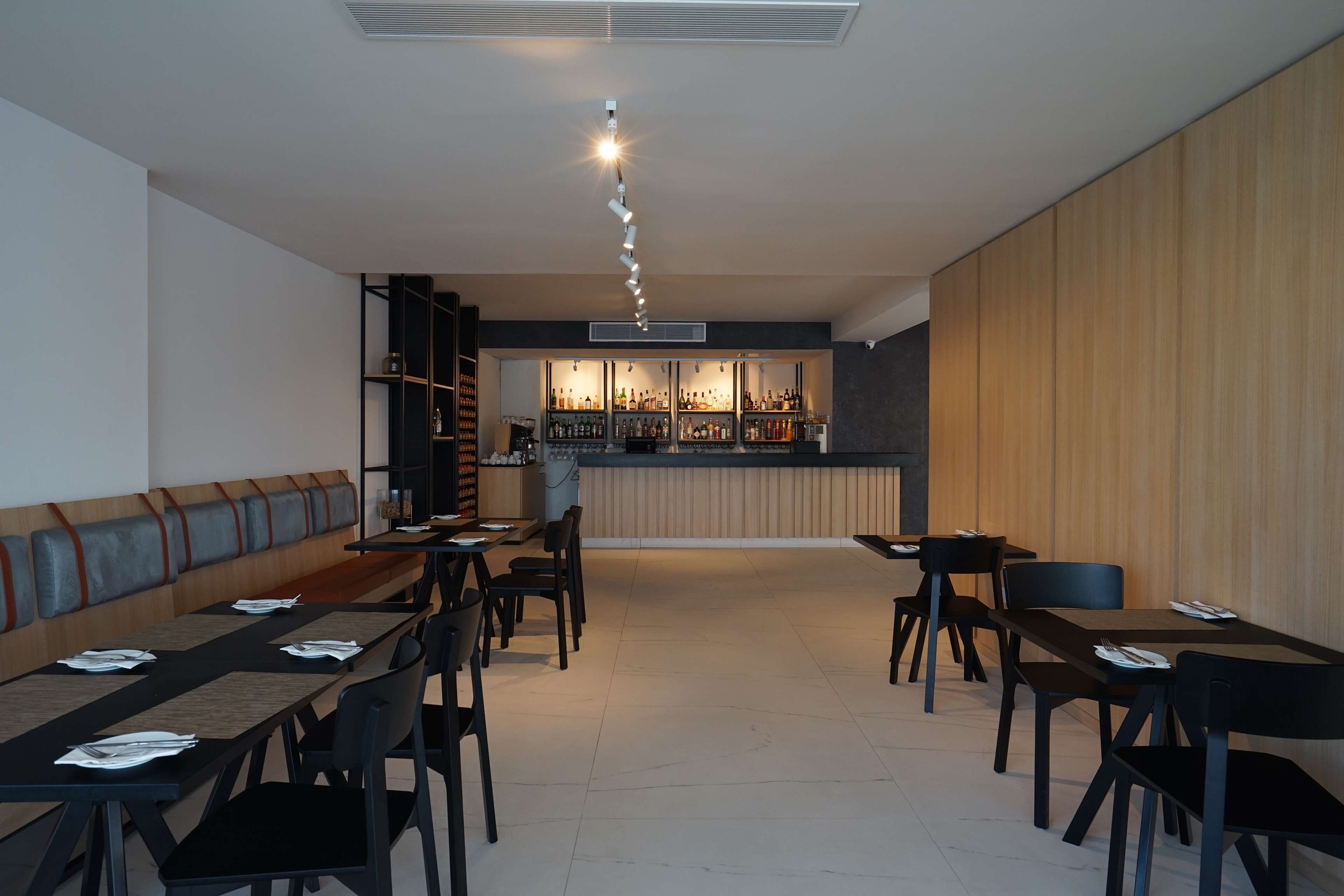 A modern restaurant with a long counter, black chairs, and a bar area in the background.