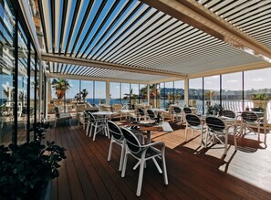 Un espacio al aire libre techado con mesas y sillas, ventanas amplias que ofrecen vistas al mar y al paisaje urbano.