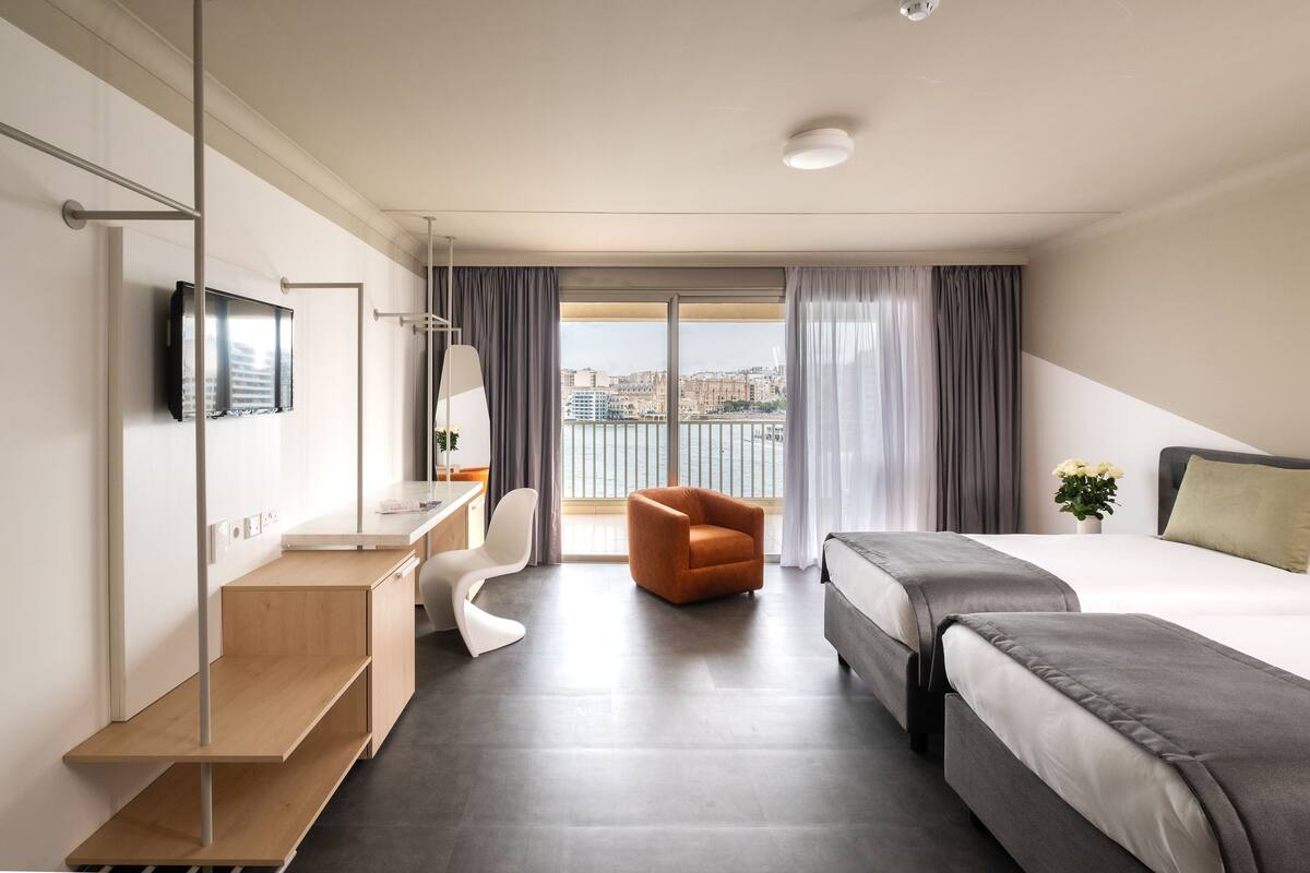 Habitación de hotel con cama, escritorio con silla, televisor y vistas a un paisaje urbano a través de amplias ventanas.