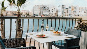 A table set with plates of food and wine glasses, overlooking a body of water with buildings in the background. - Cavalieri Hotel Malta, a member of Radisson Individuals (St. Julian's)