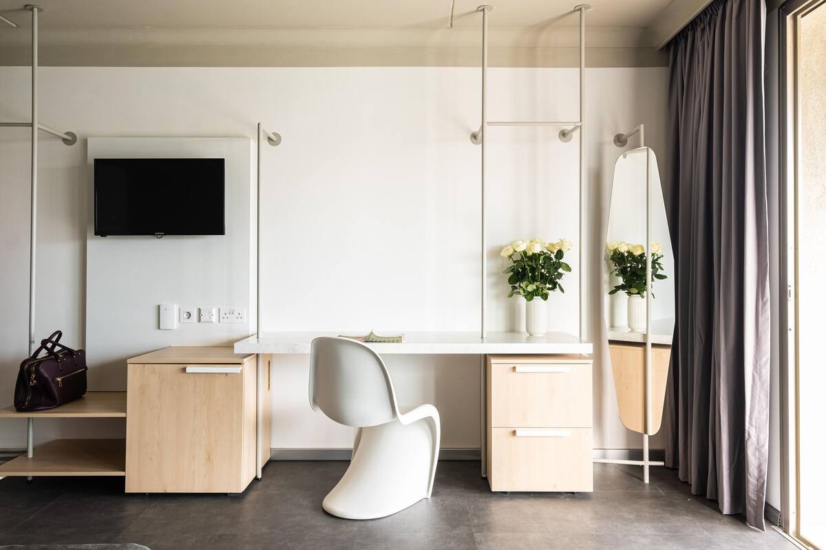 Una habitación de hotel moderna con un escritorio de madera, una silla blanca, un televisor de pantalla plana, un espejo y una ventana con cortinas.