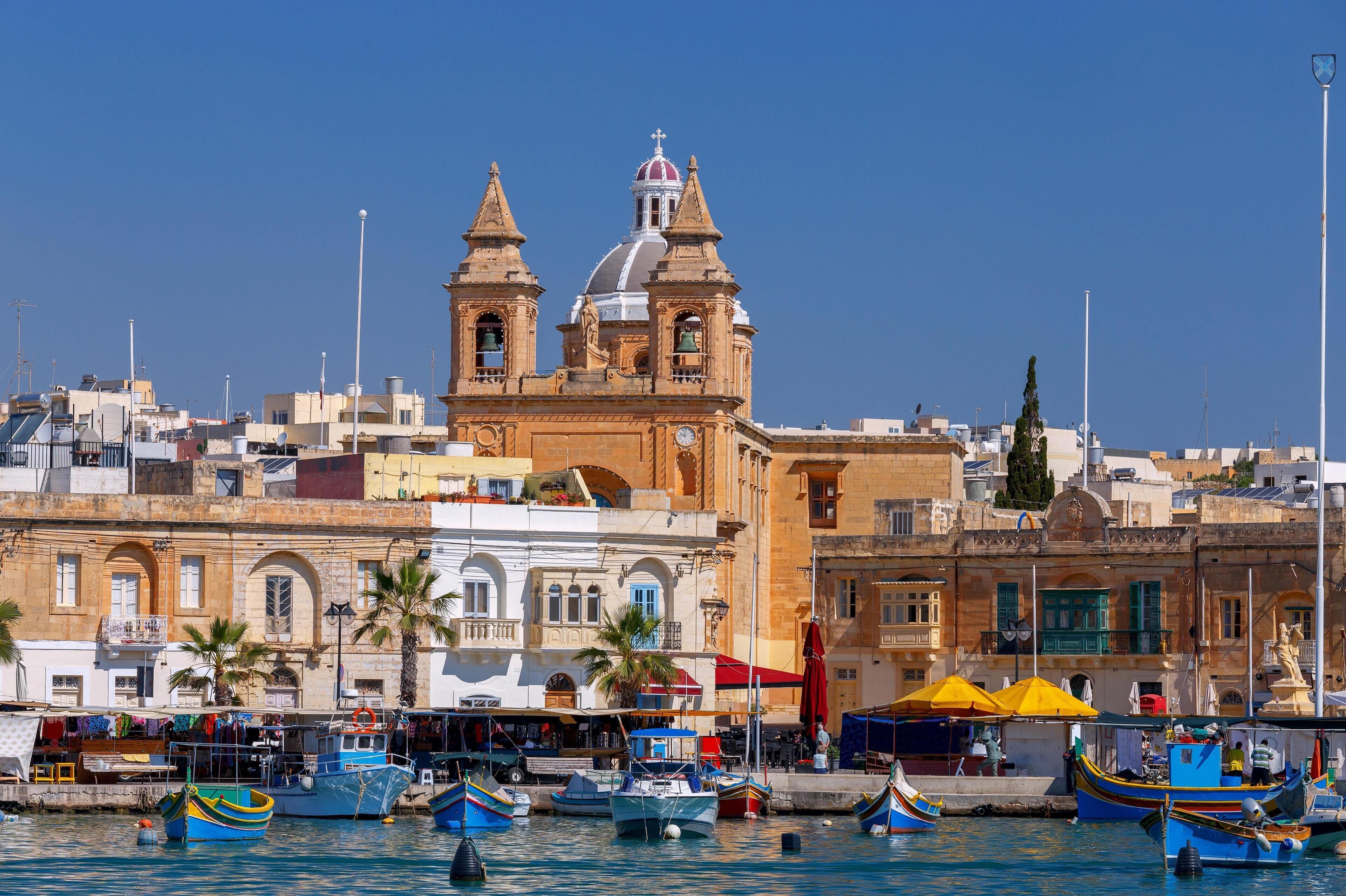 Un pueblo costero con una iglesia destacada que presenta dos torres y un campanario, un puerto deportivo con barcos de colores, y una combinación de edificios tradicionales y modernos.