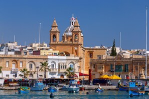 A coastal town with a prominent church featuring two towers and a bell tower, a marina with colorful boats, and a mix of traditional and modern buildings. - Cavalieri Hotel Malta, a member of Radisson Individuals (St. Julian's)