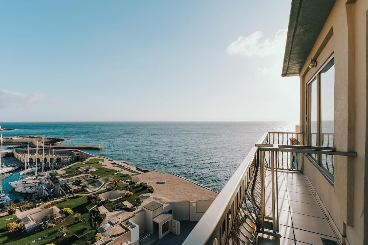 Un edificio alto con balcón que da a un puerto deportivo con yates y una playa.