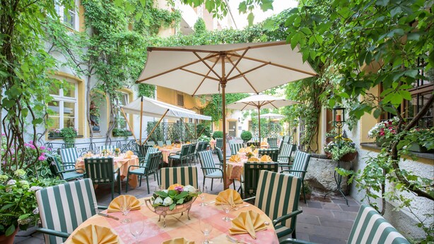 An outdoor café with tables and chairs, surrounded by greenery and a building. - Romantik Hotel zur Post (Fuerstenfeldbruck)