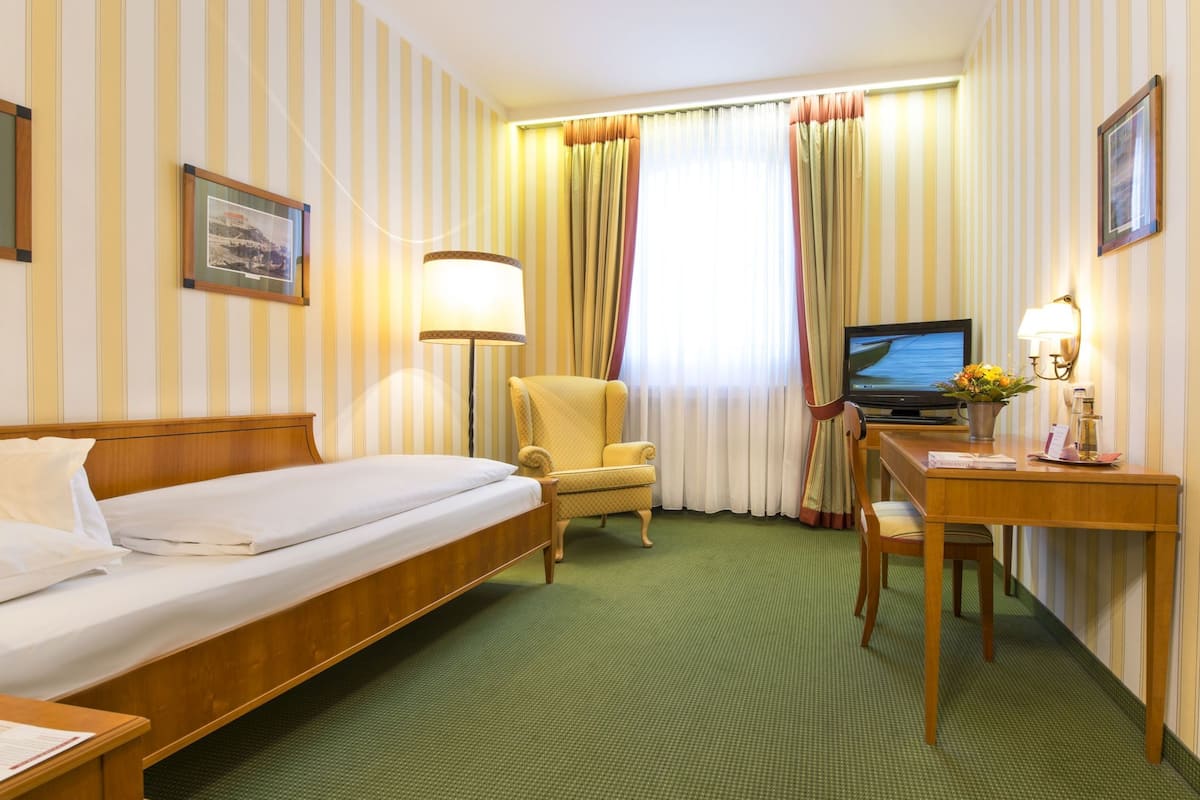 A hotel room with a single bed, a desk with a chair, a television, and a lamp.