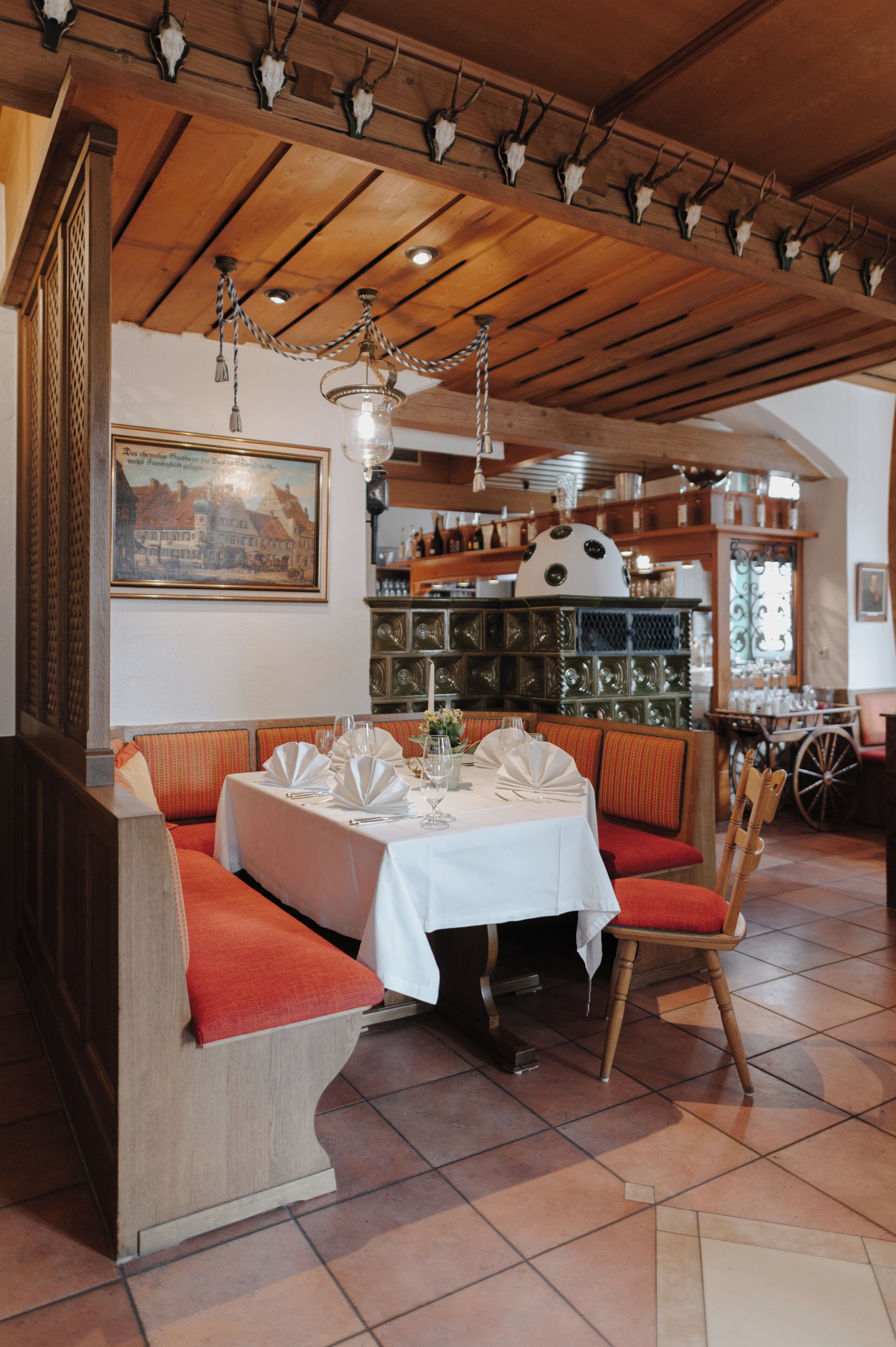 A restaurant with wooden beams, a chandelier, and a dining area with red cushioned benches and white tablecloths.