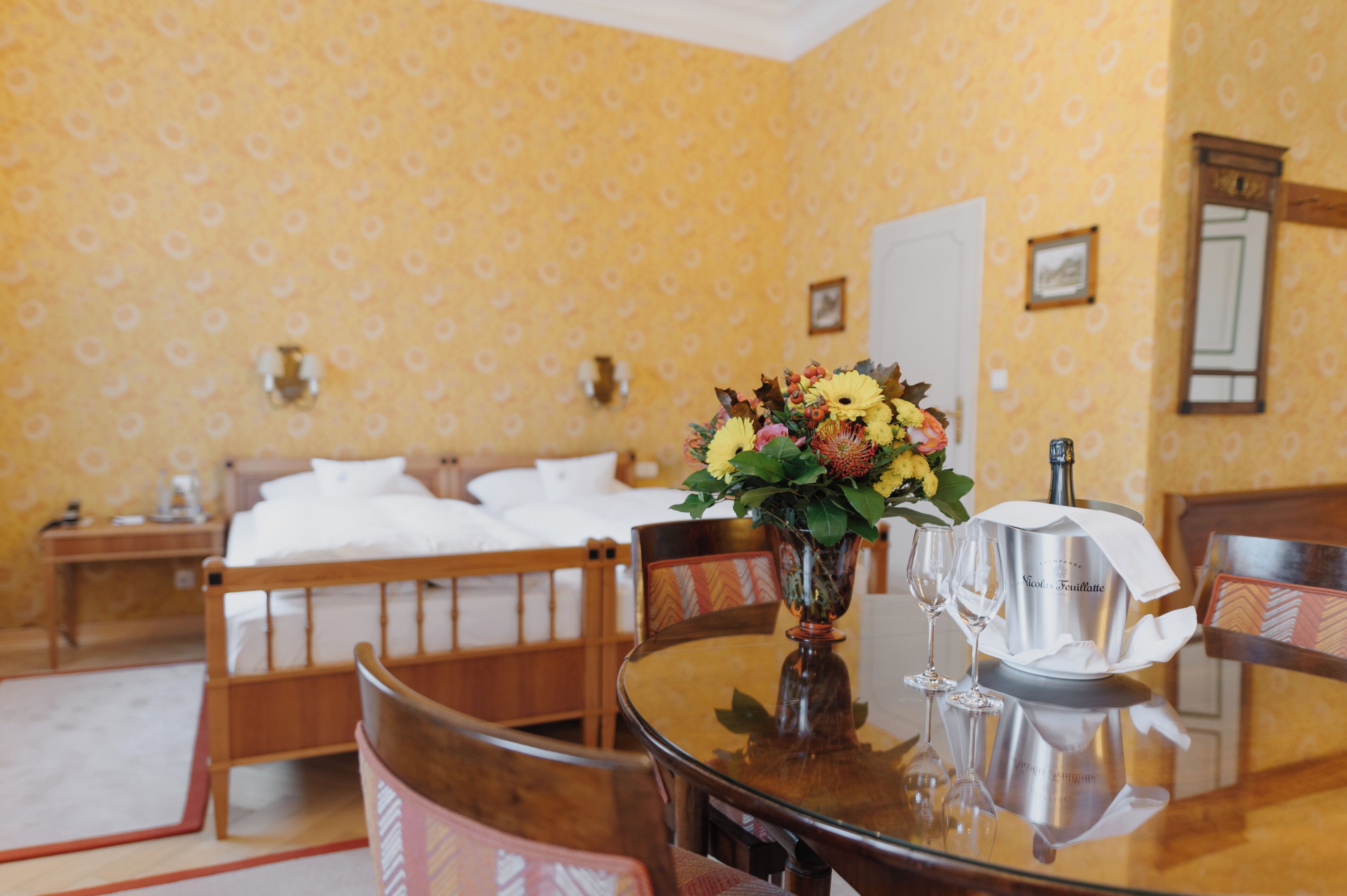 A hotel room with a bed, a round table with a vase of flowers, and a bottle of champagne.