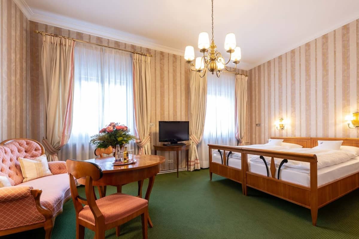 A hotel room with a bed, a chair, a small table with a vase of flowers, a television, and a chandelier.