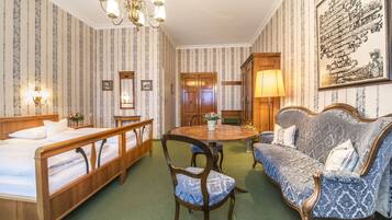 A hotel room with a bed, a wooden chair, a sofa, a dining table, and a chandelier.