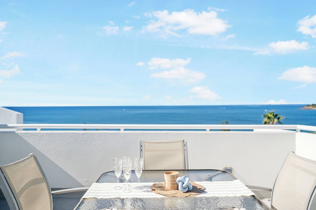 A table with a view of the ocean, a chair, and a small table with a cup and glasses.