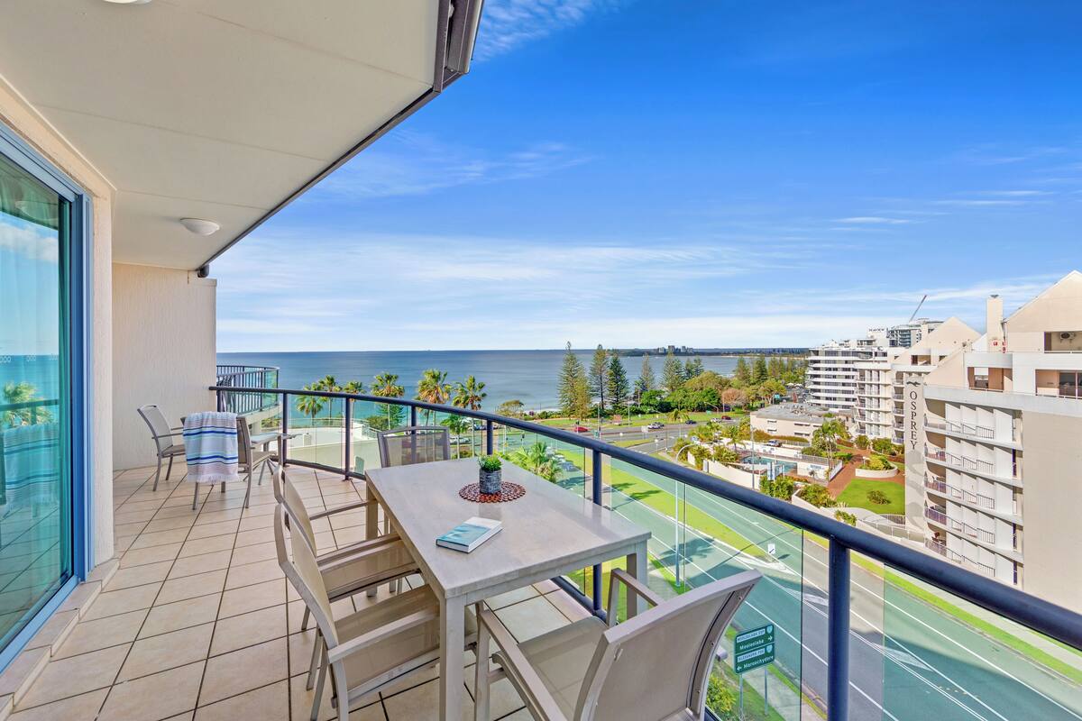 A balcony with a table and chairs, overlooking a road and buildings, with a view of the sea.