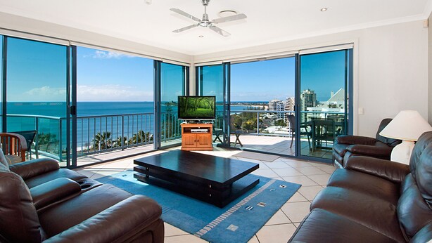 A spacious living room with a large TV, leather sofas, and a balcony with a city view. - Alexandra on the Pacific (Alexandra Headland)