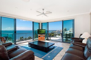 A spacious living room with a large TV, leather sofas, and a balcony with a city view. - Alexandra on the Pacific (Alexandra Headland)