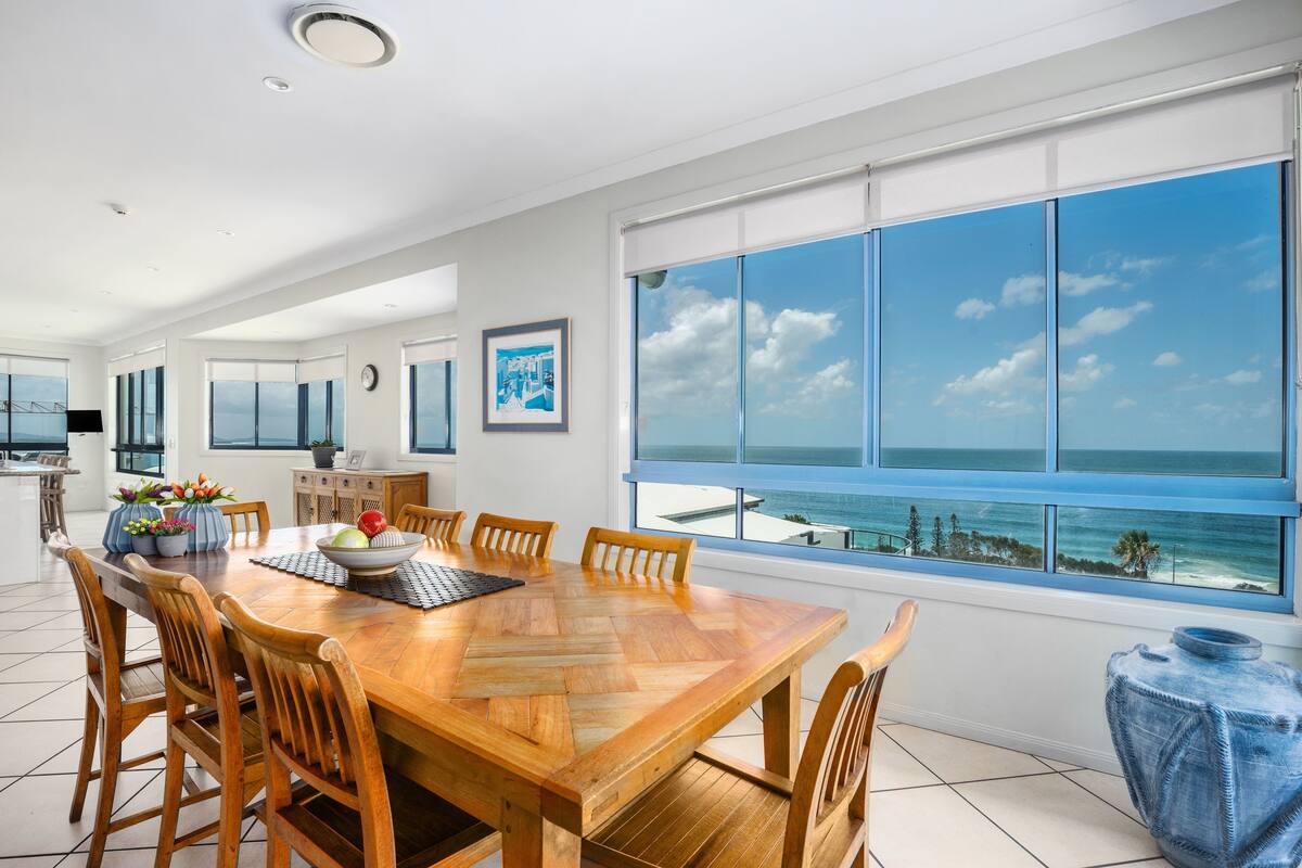 A spacious dining area with a large wooden table, surrounded by wooden chairs, and a view of the ocean through large windows.