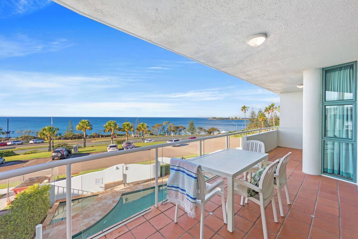 A balcony with a pool, a table, and chairs, overlooking a road and the ocean.