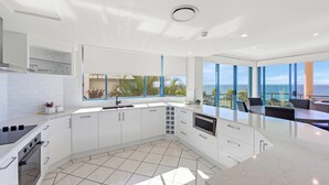 A modern kitchen with white cabinetry, a large island, and a view of the ocean. - Alexandra on the Pacific (Alexandra Headland)