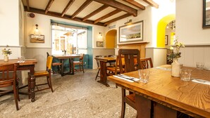 A dining area with wooden tables and chairs, a stone floor, and a view of the street through an open door. - The West Bay Hotel (Bridport)