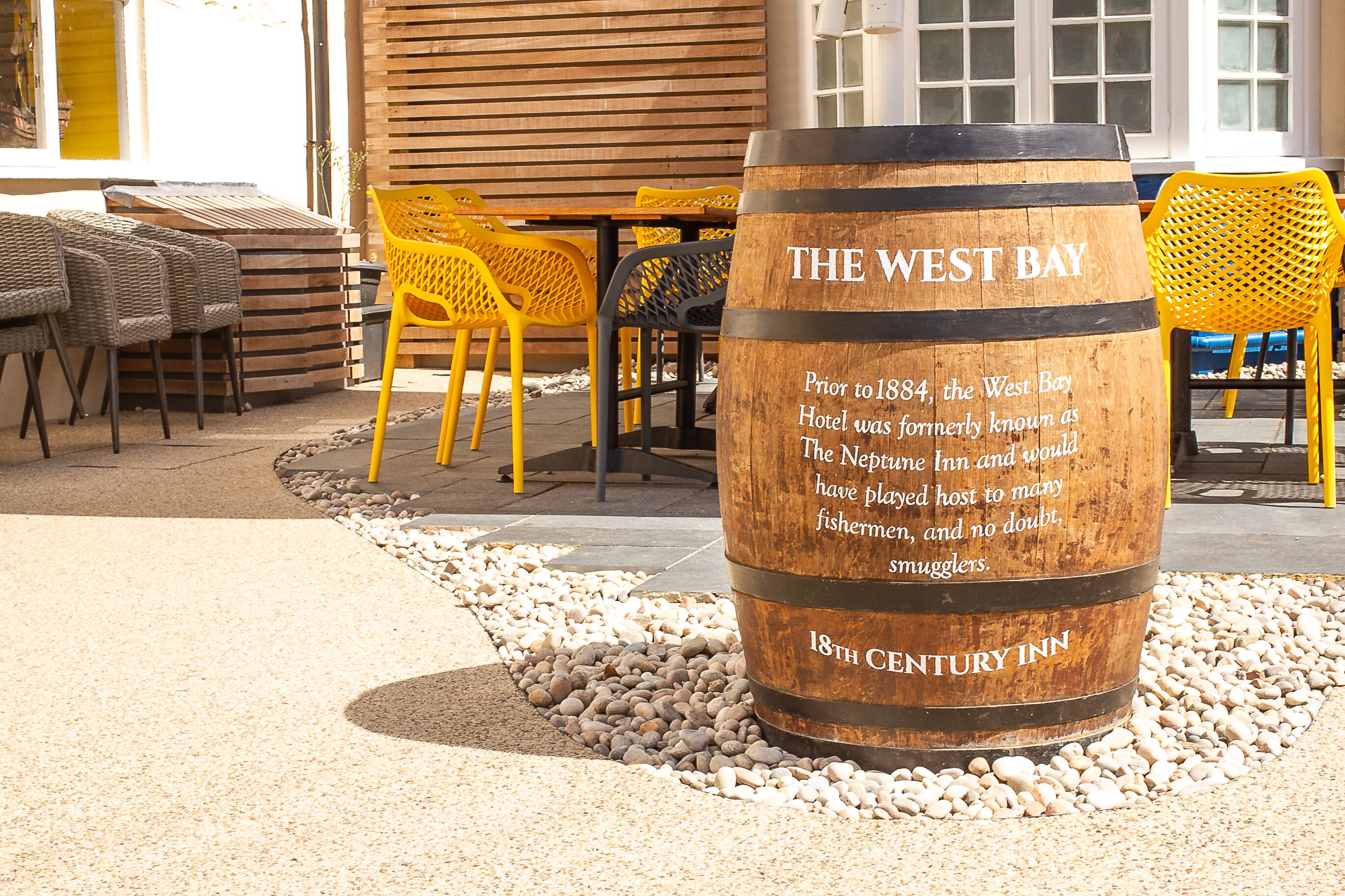 Un barril de madera con texto sobre la historia del Hotel West Bay, rodeado de mobiliario de exterior.
