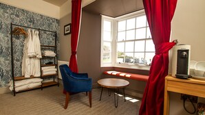 A room with a blue chair, a wooden table, a radiator, a window with red curtains, and a shelf with towels. - The West Bay Hotel (Bridport)