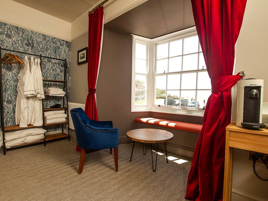 Una habitación con una silla azul, una mesa de madera, un radiador, una ventana con cortinas rojas y un estante con toallas.