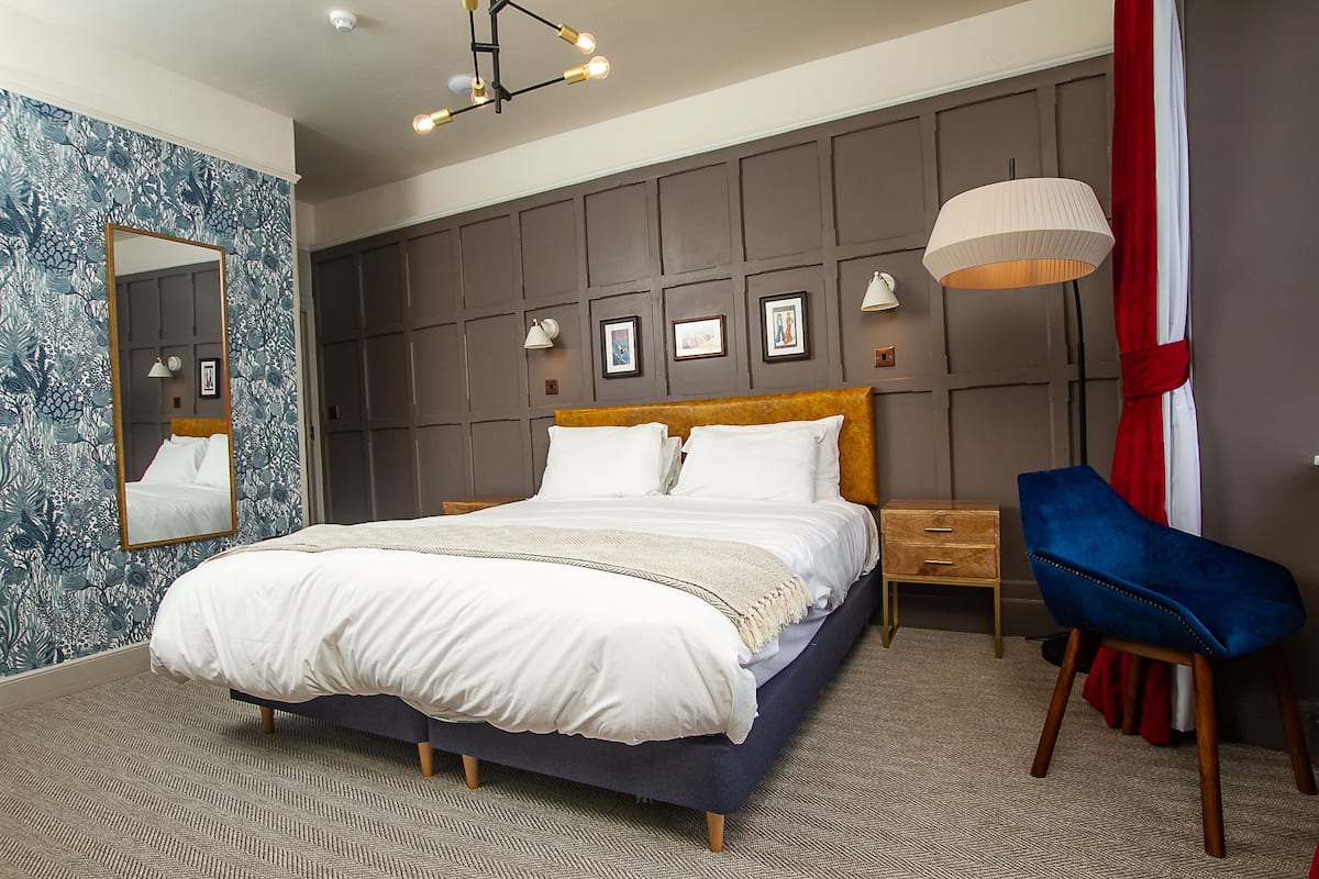 A bedroom with a large bed, a wooden nightstand, a blue chair, a mirror, and a wall with framed pictures.
