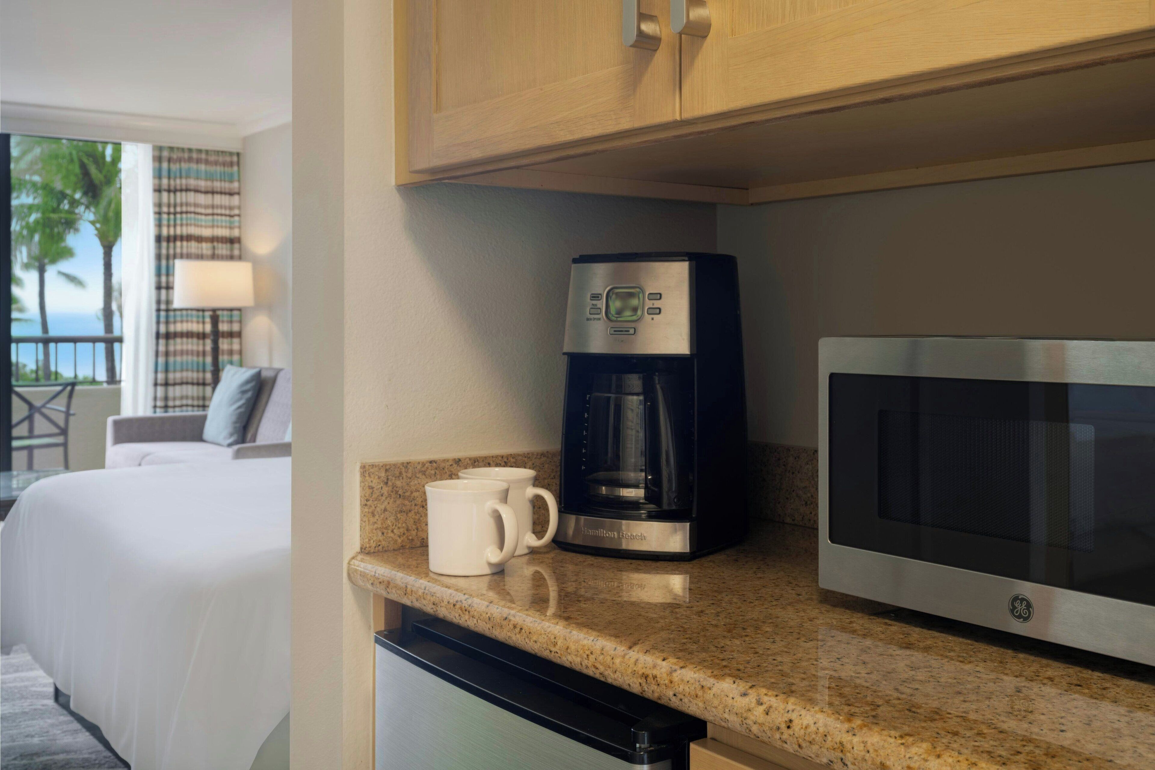 Une cuisine d’hôtel équipée d’une machine à café et d’un four à micro-ondes, ainsi qu’une chambre avec un lit donnant sur des palmiers.