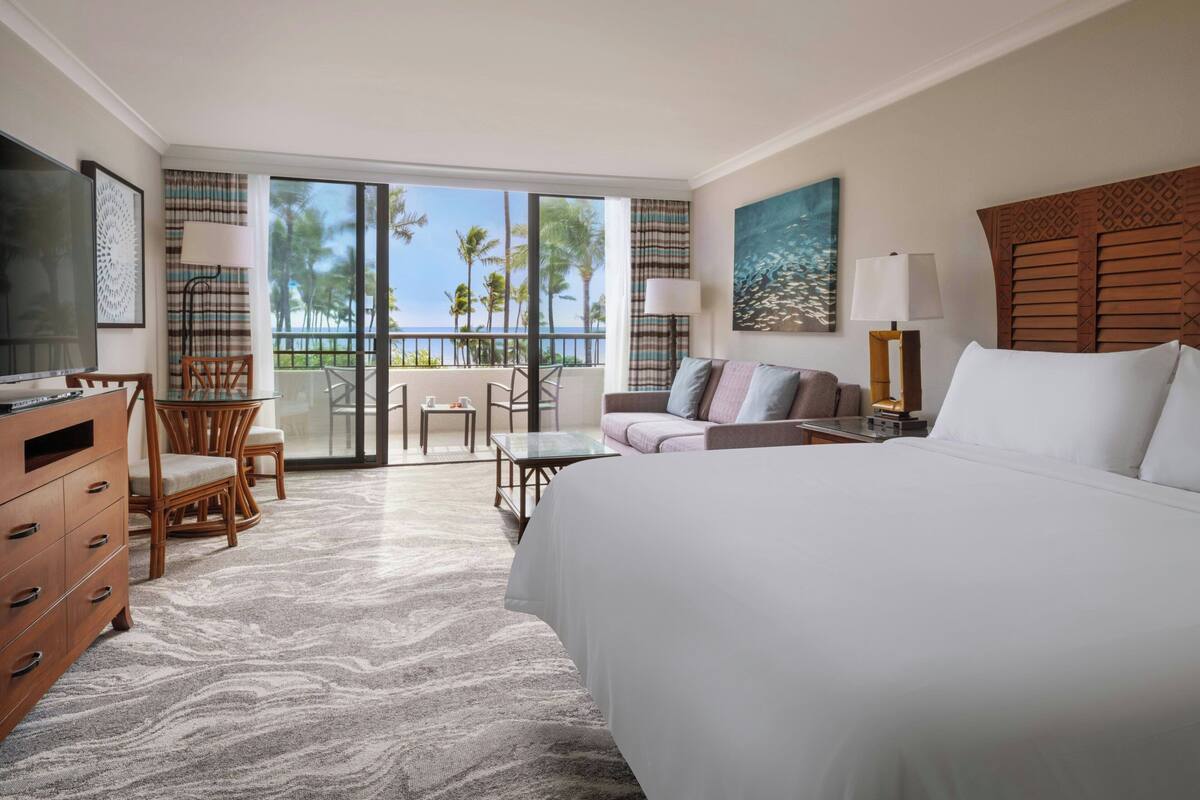 A hotel room with a large bed, a TV, a desk, and a balcony with a view of palm trees.