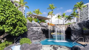 A pool with a waterfall, surrounded by rocks and palm trees, with a building in the background.