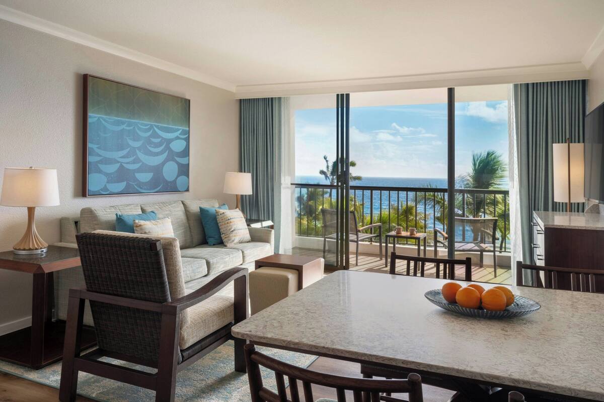 Een hotelkamer met een balkon, een eethoek en een groot raam met uitzicht op de oceaan.