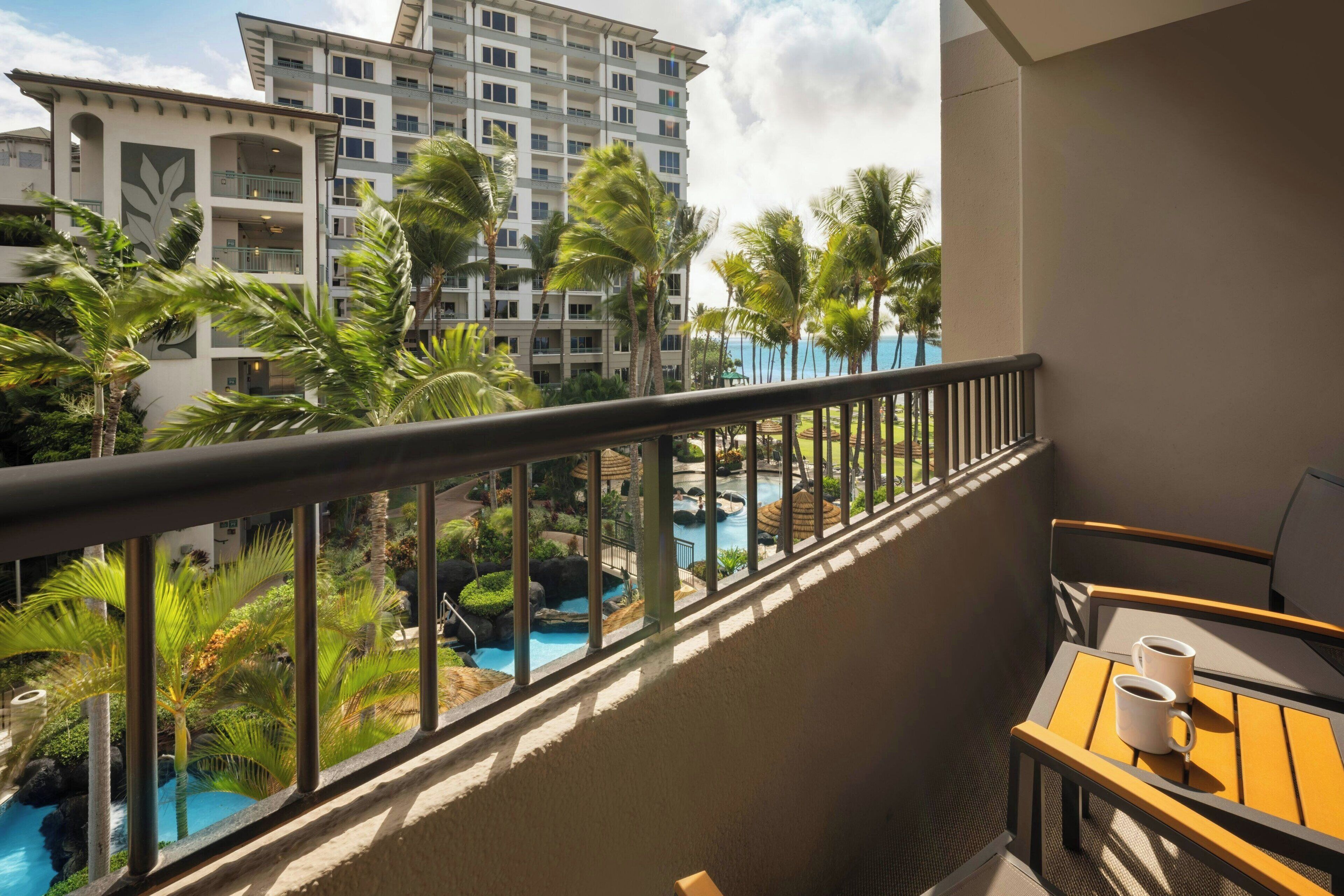 A balcony with a view of a swimming pool, palm trees, and a hotel building.