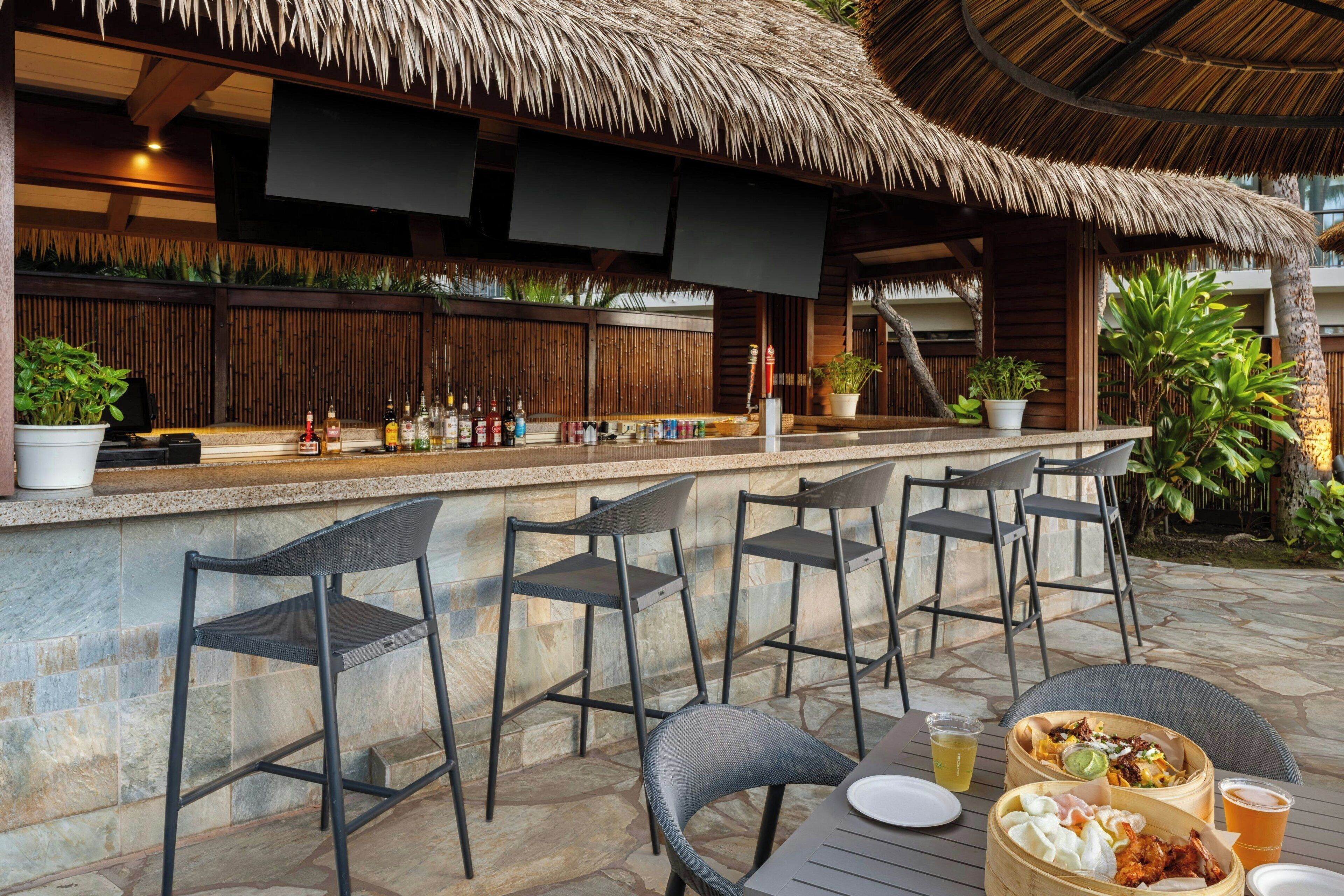 An outdoor bar area with a thatched roof, stone walls, and potted plants.