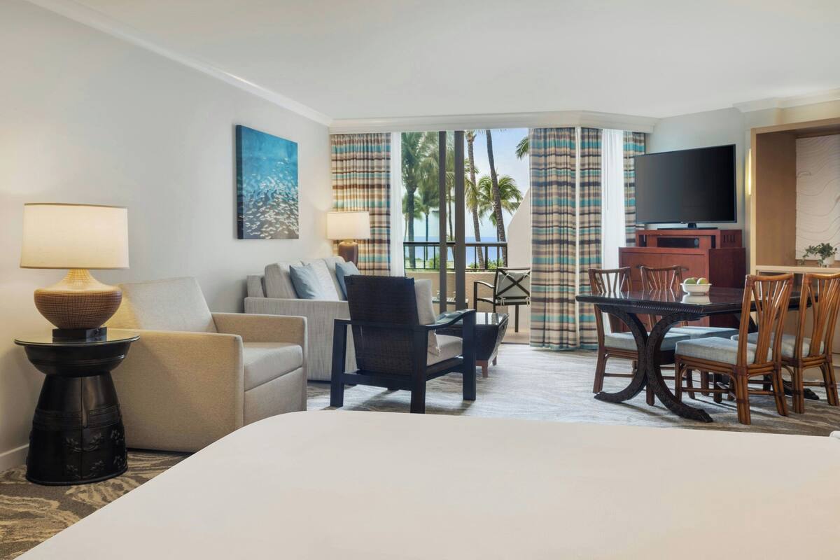 A hotel room with a large bed, a sofa, a dining table with chairs, a TV, and a balcony with curtains.