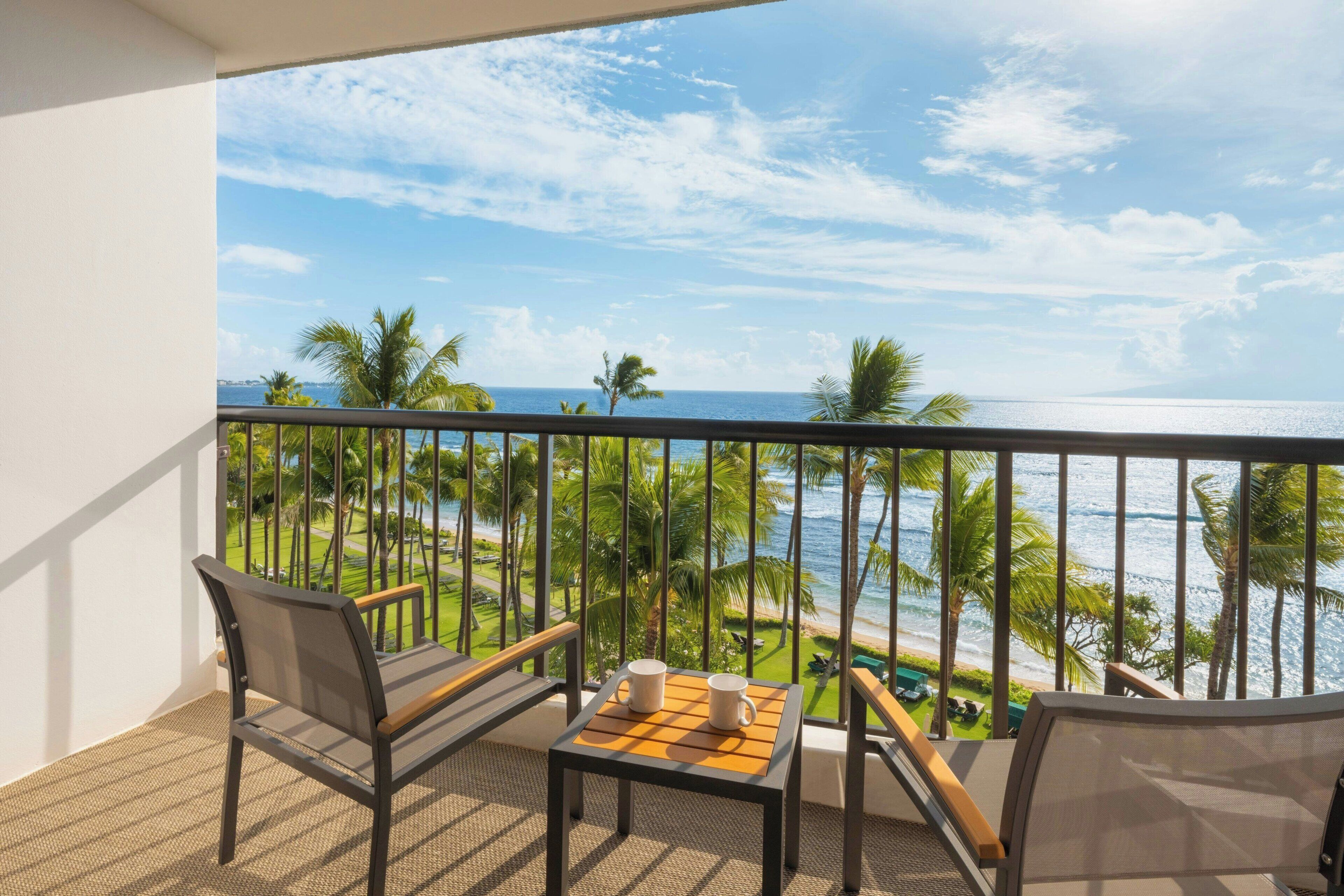 A balcony with a view of the ocean, two chairs, a small table with two cups, and a pool in the background.